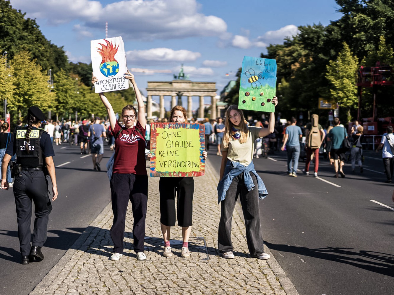 Drei junge Frauen halten Protestplakate in die Luft. Sie stehen vor dem Brandenburger-Tor bei einer Klimademo. Die Schilder sind selbstgemalt mit Aufschriften wie: Ohne Glaube – keine Veränderung.